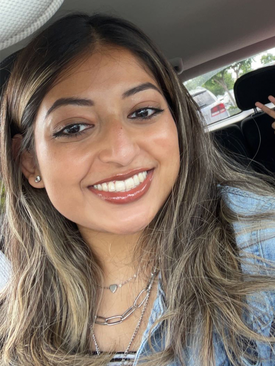 A woman with long, wavy brown hair smiles at the camera while sitting in a car, wearing a denim jacket and layered necklaces.