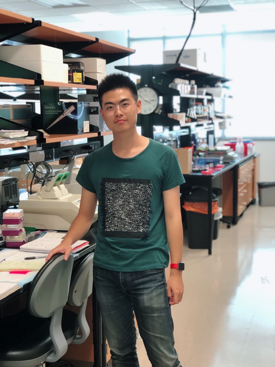 A person stands next to a lab bench in a bright laboratory, surrounded by scientific equipment, notebooks, and shelves with supplies.