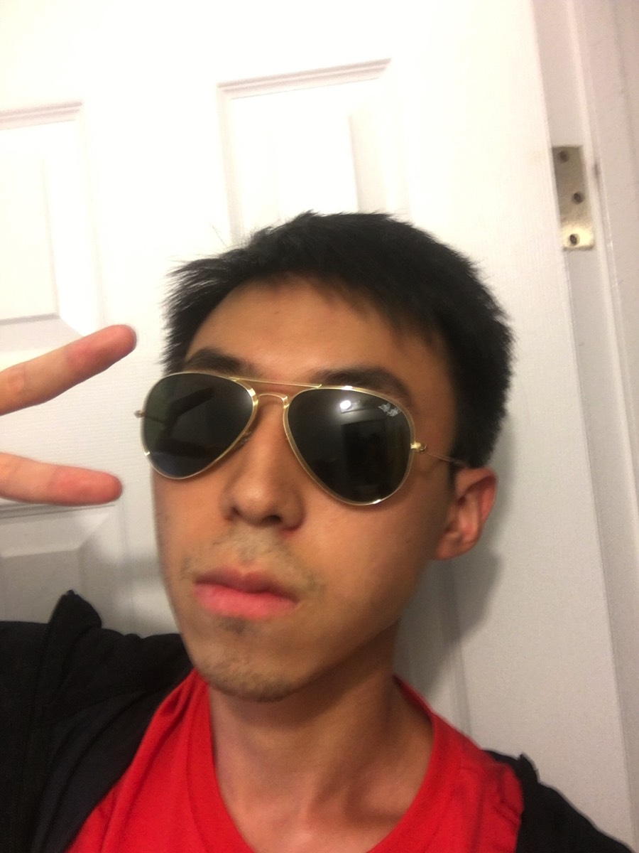 A person wearing aviator sunglasses and a red shirt stands indoors, making a peace sign with their hand in front of a white door.