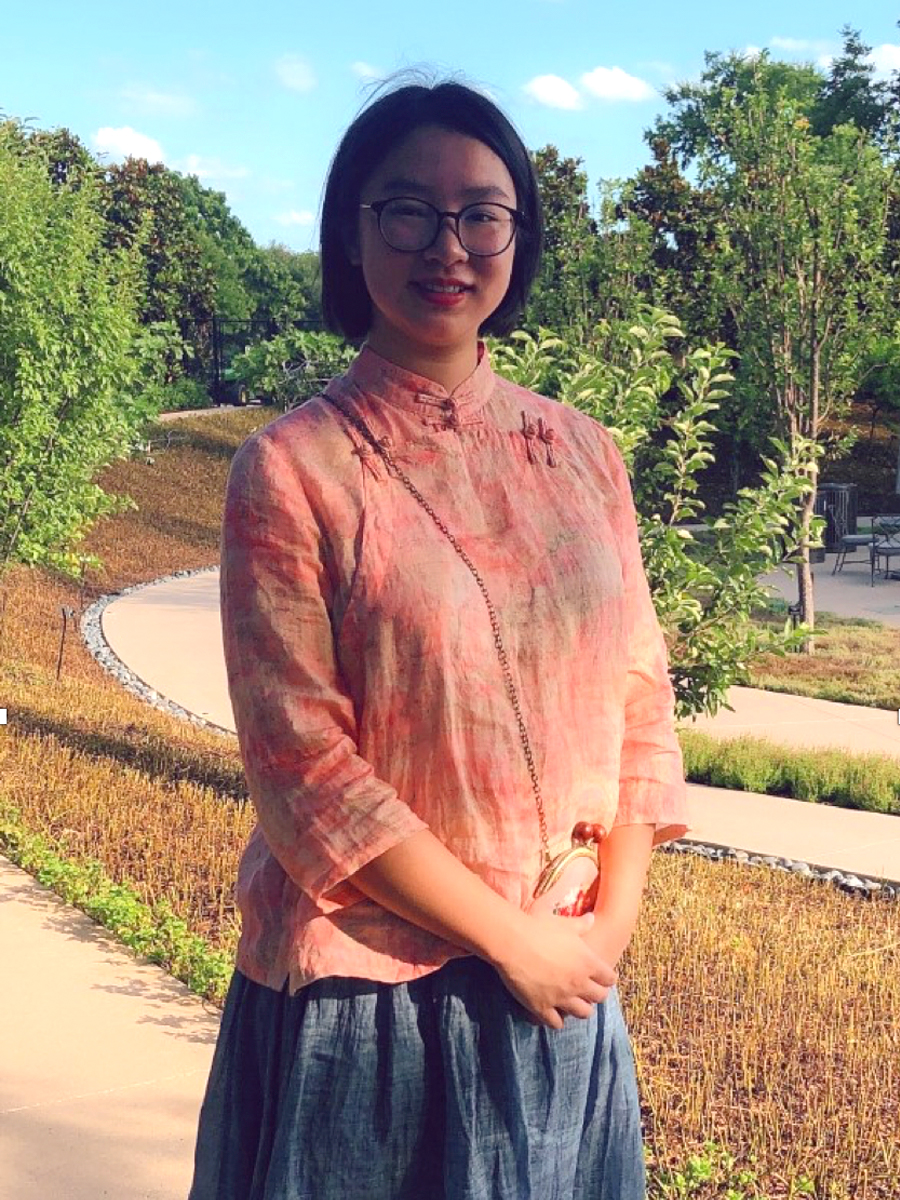 A woman wearing glasses and a pink top stands outdoors on a sunny day, with greenery and a winding path in the background.