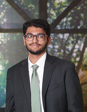 A man wearing glasses, a dark suit, white shirt, and green tie stands in front of a blurred backdrop featuring window panes and greenery.
